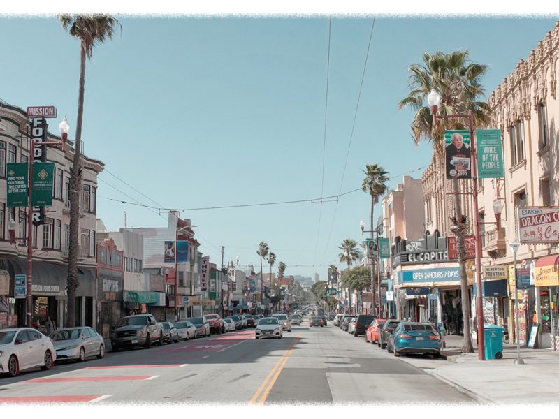 A busy urban street with parked cars and storefronts, palm trees lining the road. Several business signs and advertisements are visible, with pedestrians along the sidewalks.