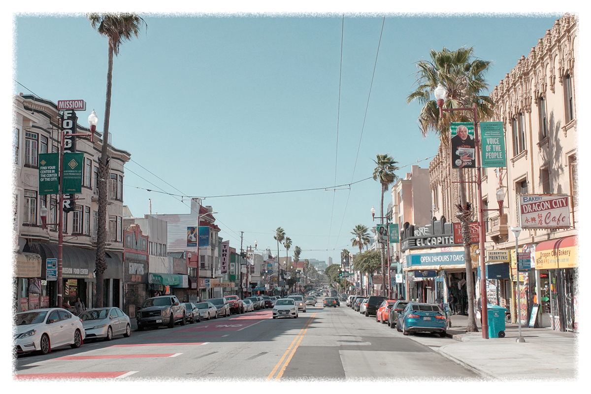 A busy urban street with parked cars and storefronts, palm trees lining the road. Several business signs and advertisements are visible, with pedestrians along the sidewalks.