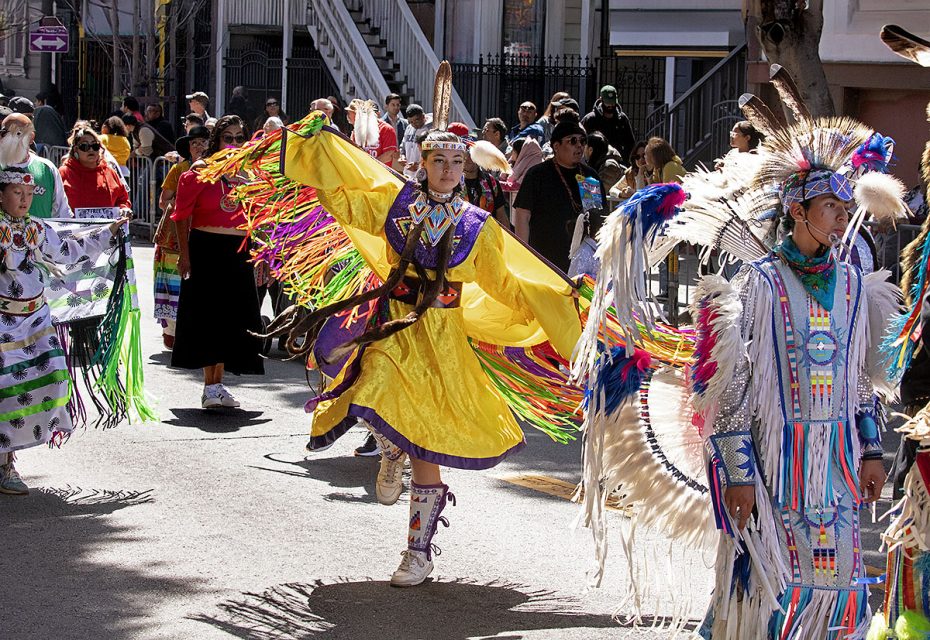 A group of people in colorful, traditional Native American attire participate in a street parade, showcasing vibrant feathers and intricate beadwork.