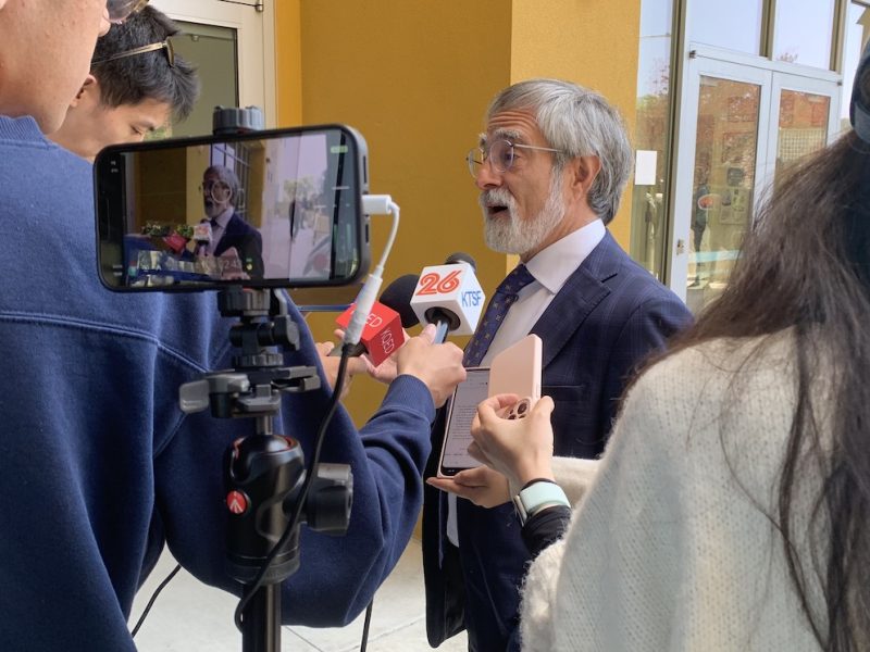 A man in a suit speaks to multiple reporters holding microphones and recording devices in front of a building. A camera on a tripod is also capturing the scene, as Peskin addresses the media with poised confidence.