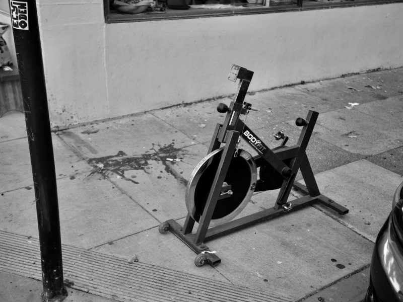 A stationary exercise bike is placed on a sidewalk next to a metal pole and a street curb.