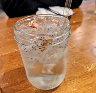 A clear glass jar filled with a chilled drink containing ice cubes and chia seeds sits on a wooden table, with a straw and a napkin in the background.