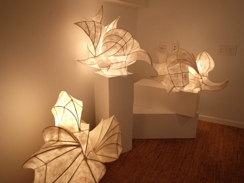 Three abstract, illuminated paper sculptures on white pedestals are displayed in a well-lit gallery with cork flooring.
