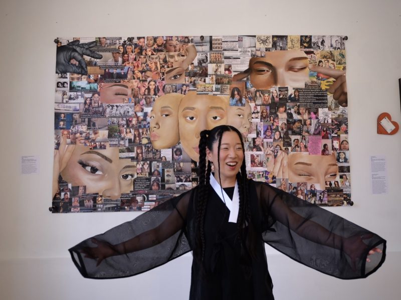 A person with braided hair and transparent sleeves stands smiling in front of a collage artwork featuring various eyes and faces.