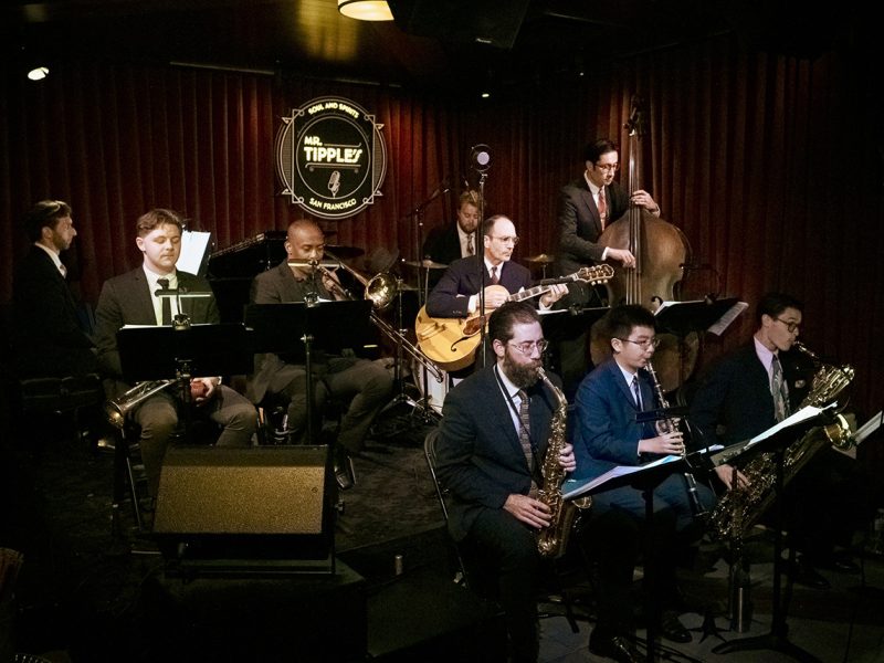 A jazz band performs on stage at Mr. Tipple's, featuring various musicians playing brass, woodwind, guitar, piano, and double bass instruments. The backdrop is a maroon curtain with a logo. They'll soon play Duke Ellington classics at the CMC.