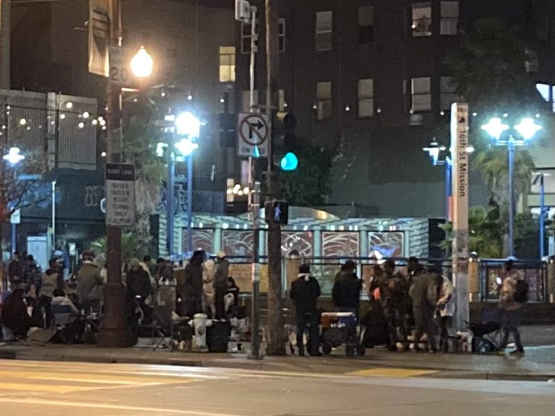 A busy street corner at night with a group of people gathered, street lights illuminated, and traffic signs visible.