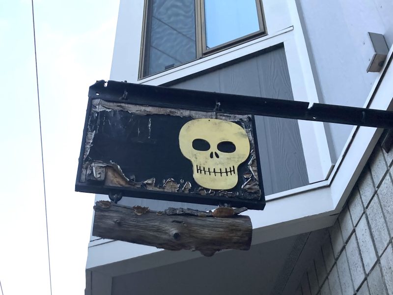 A worn-out hanging sign featuring a skull painted on a black background, affixed to a weathered wooden beam outside a building.