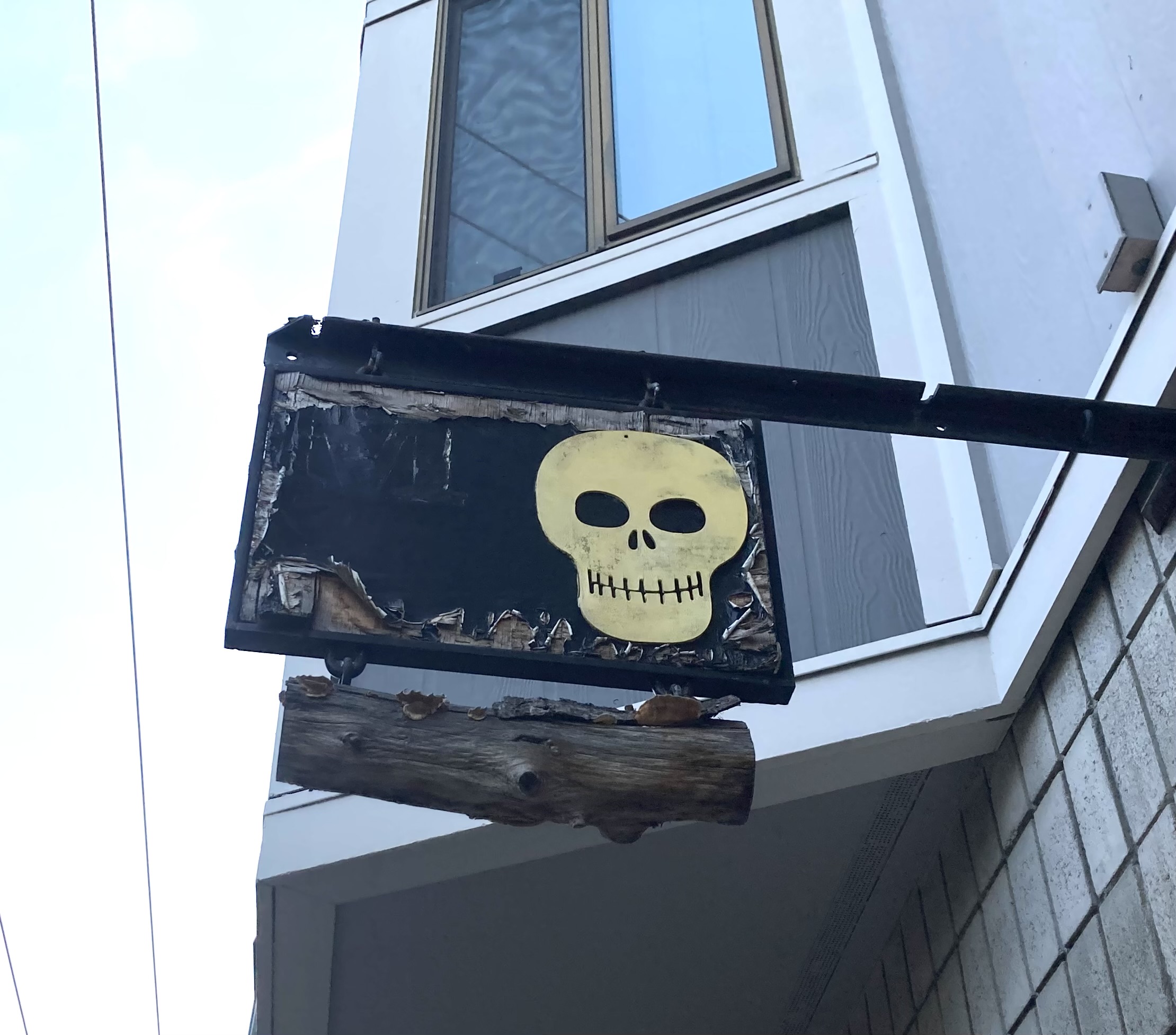A worn-out hanging sign featuring a skull painted on a black background, affixed to a weathered wooden beam outside a building.