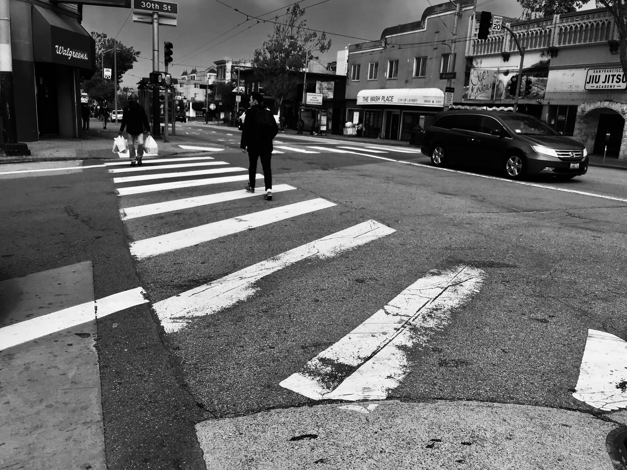 Black and white image of people crossing a street using a zebra crossing in an urban area with cars, buildings, and a traffic light visible in the background.