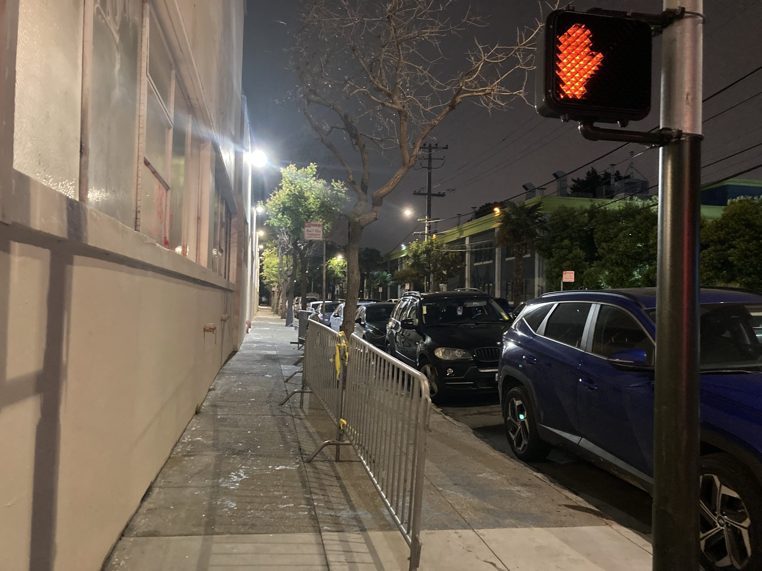 Nighttime street scene with a "Don't Walk" signal illuminated. The sidewalk is lined with parked cars and a barricade, and a leafless tree is visible in the background.