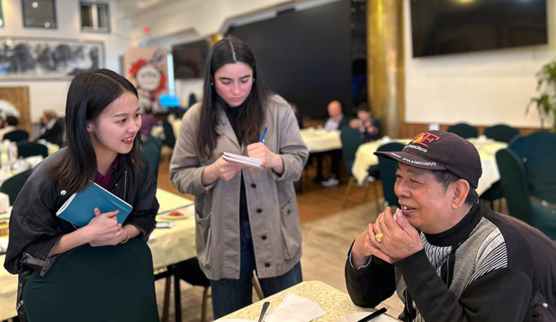 Two women taking notes while listening to an elderly man speaking at a community hall event.