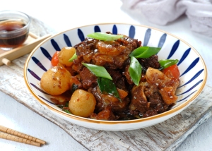 A bowl of braised oxtail mixed with vegetables and garnished with chopped green onions, accompanied by a small dish of soy sauce and chopsticks on a white surface.