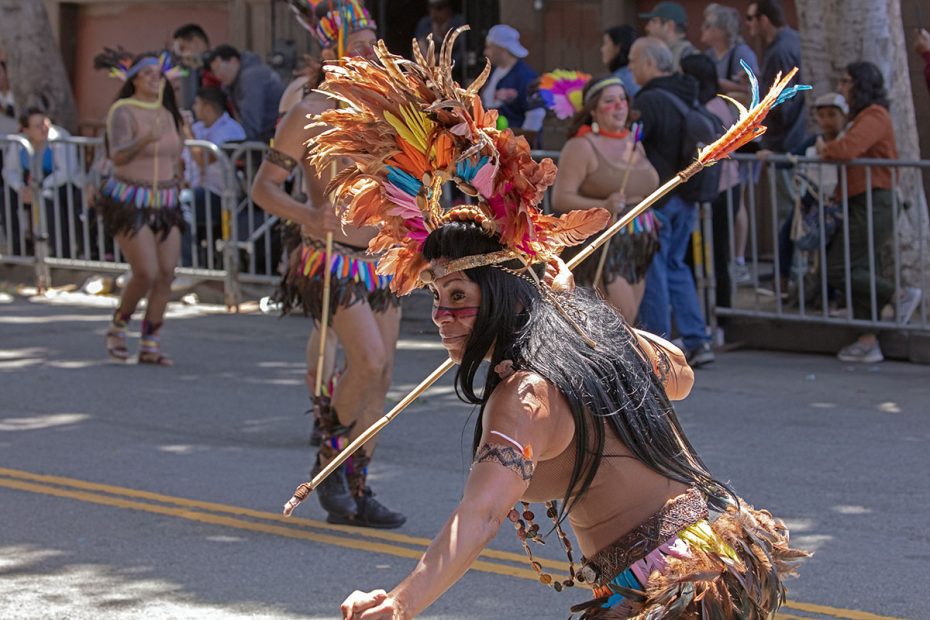 A person dressed in traditional indigenous attire with colorful feathers and face paint performs in a street parade, with onlookers watching from behind a barricade.