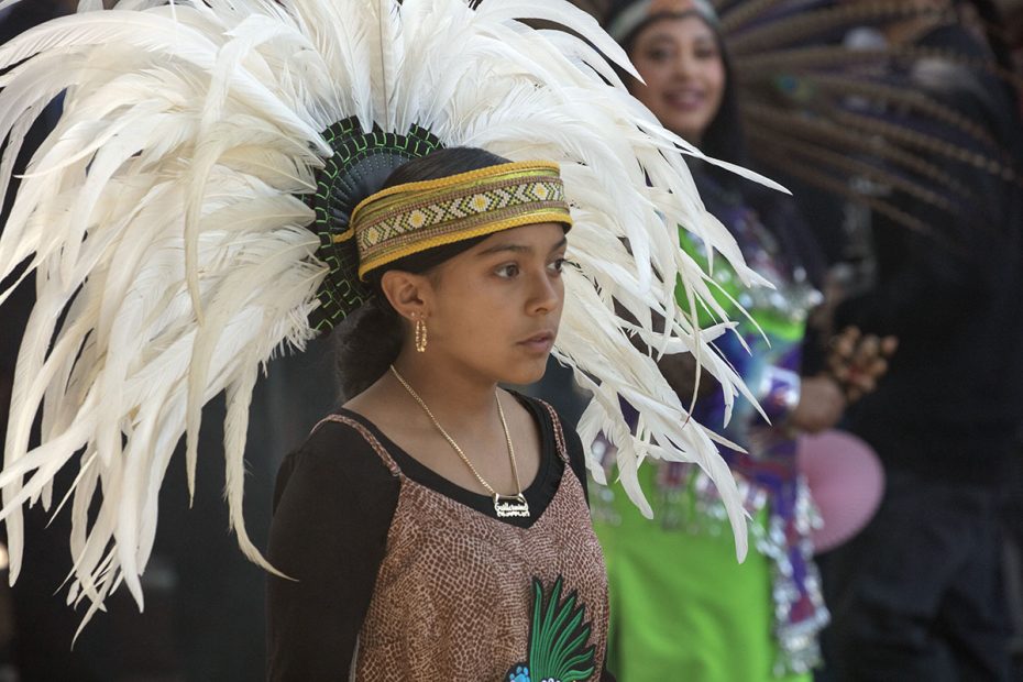 A young girl wearing a traditional feathered headdress and ornate headband participates in a cultural event. Another person dressed in colorful attire is visible in the background.
