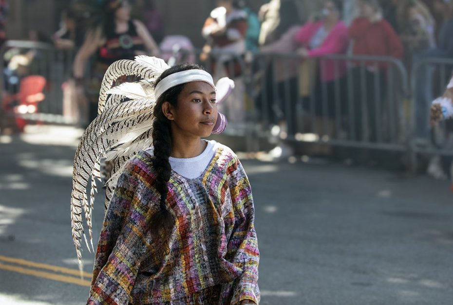 A person wearing traditional attire with a headdress of feathers participates in a parade.