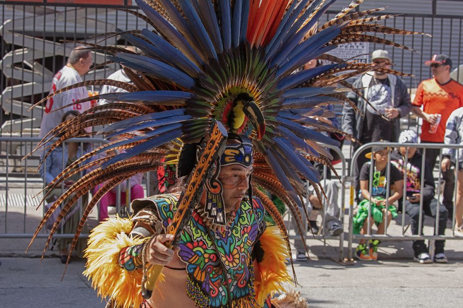Person in vibrant, traditional attire with colorful feathered headdress participating in a public event, with onlookers in the background.