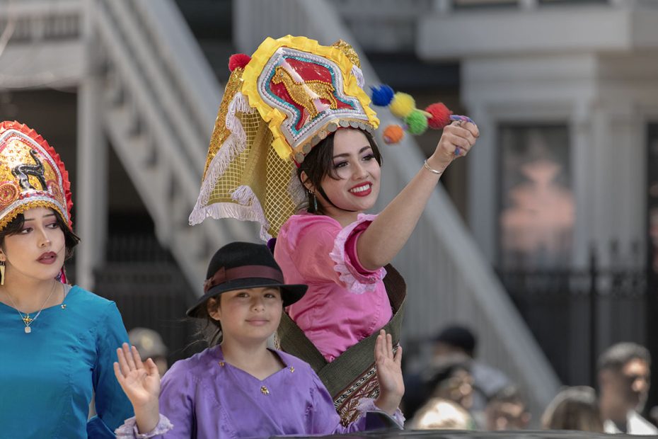 Three women in colorful traditional attire, one in pink holding a colorful tassel, participate in an outdoor cultural event, with a blurred urban background.
