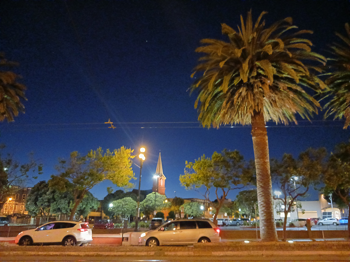Nighttime view of a city street with palm trees, parked cars, and a church steeple illuminated against a dark blue sky.