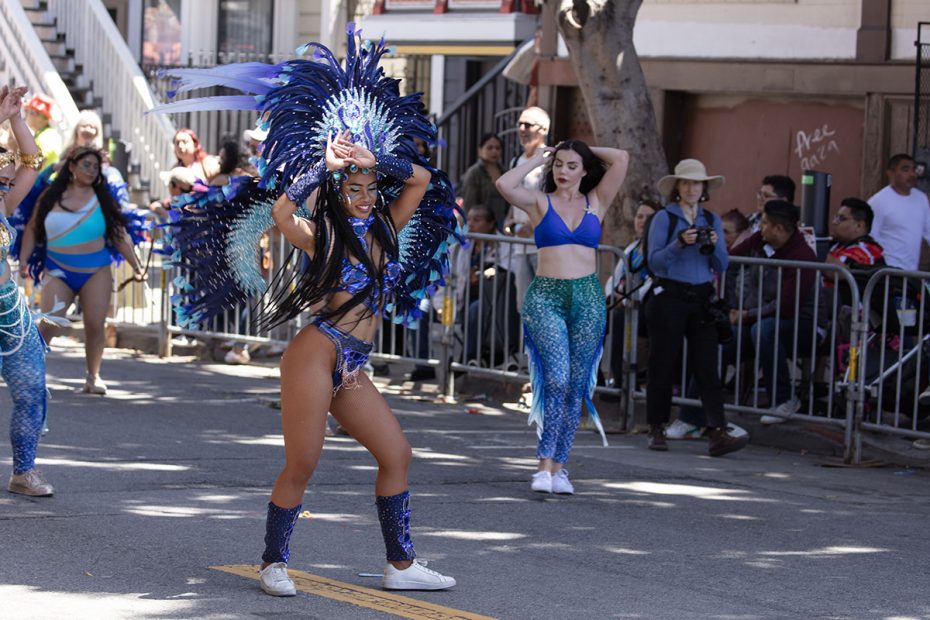 A performer in a vibrant blue and purple feathered costume dances in a street parade, while onlookers watch from behind barriers. Other performers and spectators are visible in the background.