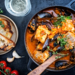 A pot of seafood stew with mussels and white fish is garnished with herbs. A plate with bread slices, whole garlic cloves, cherry tomatoes, and a glass of white wine are adjacent.