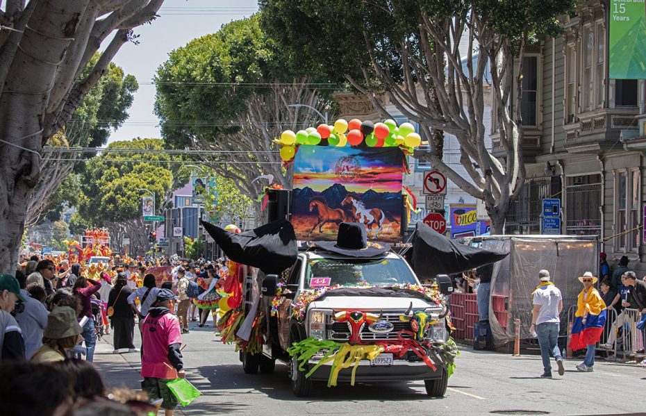 A decorated truck with large black wings and colorful balloons participates in a street parade, surrounded by spectators and trees lining the street.