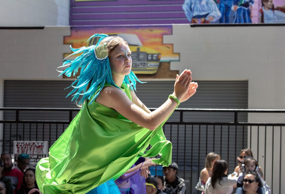 A person wearing a vibrant green costume with blue headgear performs a dance outdoors, with an audience seated in the background.