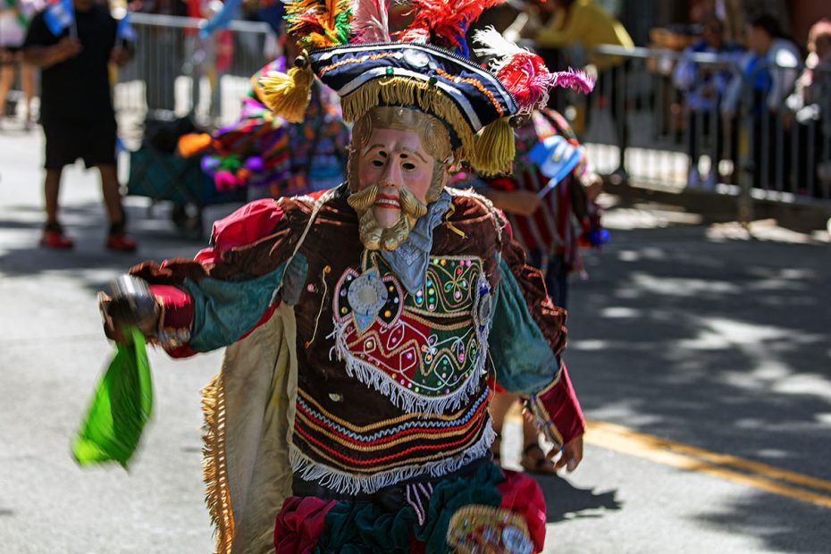 A person in a colorful, traditional costume and mask participates in a parade. The outfit includes vibrant fabrics, tassels, and a feathered headdress.
