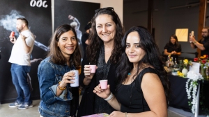 Three women holding drinks and smiling at a social event with other people in the background.