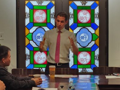Man in a suit and red tie giving a presentation at a table, with colorful stained glass windows in the background.