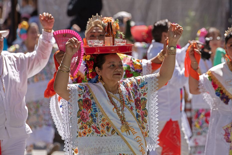 A woman in traditional attire leads a parade while balancing a red tray with drinks on her head. Other participants in colorful costumes are visible behind her.