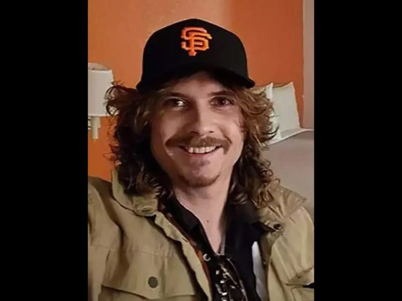 A smiling man with curly hair wearing a san francisco giants baseball cap and a khaki jacket, seated in a room with an orange wall.