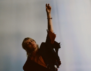 Person in an orange jacket and black pants reaching upwards against a muted blue background.