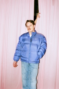 A person wearing a blue puffer jacket and light jeans stands in front of pink curtains. Another person's hand is holding the curtains open above them.