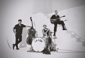 Three musicians pose with their instruments: one leans on a double bass, another sits at a drum set labeled "LA LOM," and the third is seated on stairs playing a guitar. The setting is minimalistic.