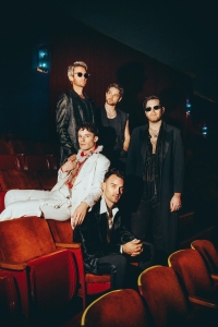 Five people dressed in formal attire are seated and standing in a dimly lit auditorium with red seats.