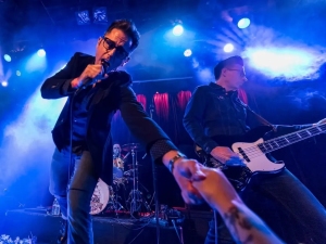 Musicians perform on stage under blue and purple lights. The lead singer with a microphone is bent forward, reaching out to touch an audience member's hand, while a guitarist plays in the background.