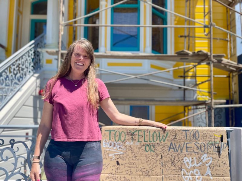 A woman in a pink shirt stands in front of a partially renovated yellow house with scaffolding. She rests her hand on a graffiti-covered barrier that reads "TOO YELLOW OR AWESOME?".
