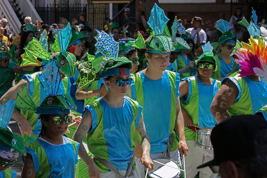 A group of people wearing blue and green costumes with aquatic-themed hats and sunglasses are participating in a parade, with some playing percussion instruments.