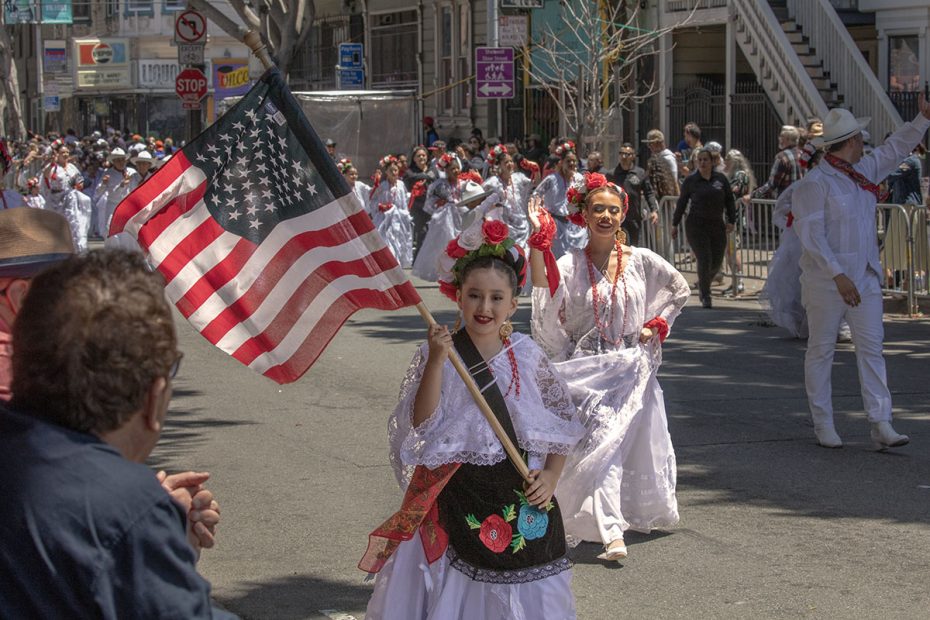 A group of people in traditional dress walk in a parade, with one person prominently holding an American flag. Several onlookers are visible in the foreground.