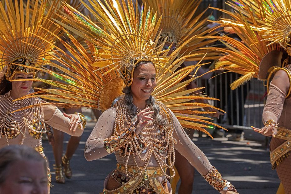 Performers in elaborate golden costumes with spiky headdresses and bead embellishments participate in a street parade. They are smiling and dancing, with a crowd and barriers visible in the background.