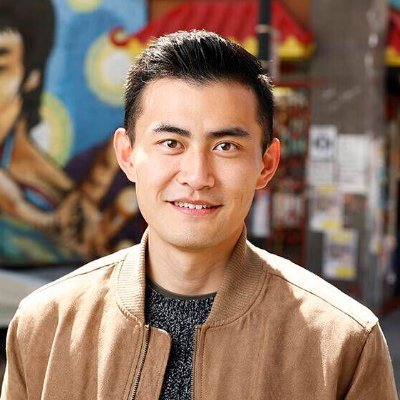 A smiling asian man in a tan jacket and grey sweater, with a colorful mural in the background.