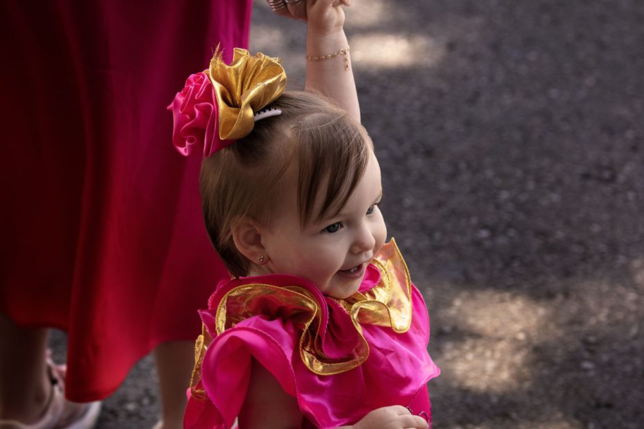 A toddler wearing a bright pink and gold outfit smiles and reaches upwards, standing outdoors on a sunlit day.