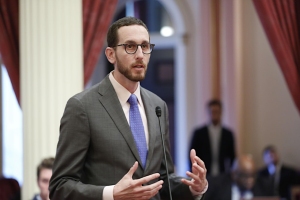 A man in a suit and glasses speaking at a podium in a room with red curtains and ornate columns.