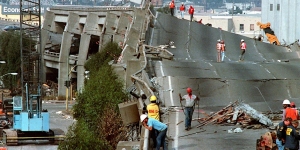 A collapsed section of a multi-lane highway with several workers in safety gear assessing the damage. Distant buildings and construction equipment are visible in the background.