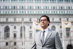 A man in a gray suit and glasses stands confidently in front of a large, ornate building with multiple windows and flags in the background.