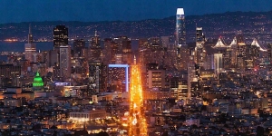 A night view of a cityscape with tall buildings and illuminated streets. A long, brightly lit street runs through the center, and hills can be seen in the background.