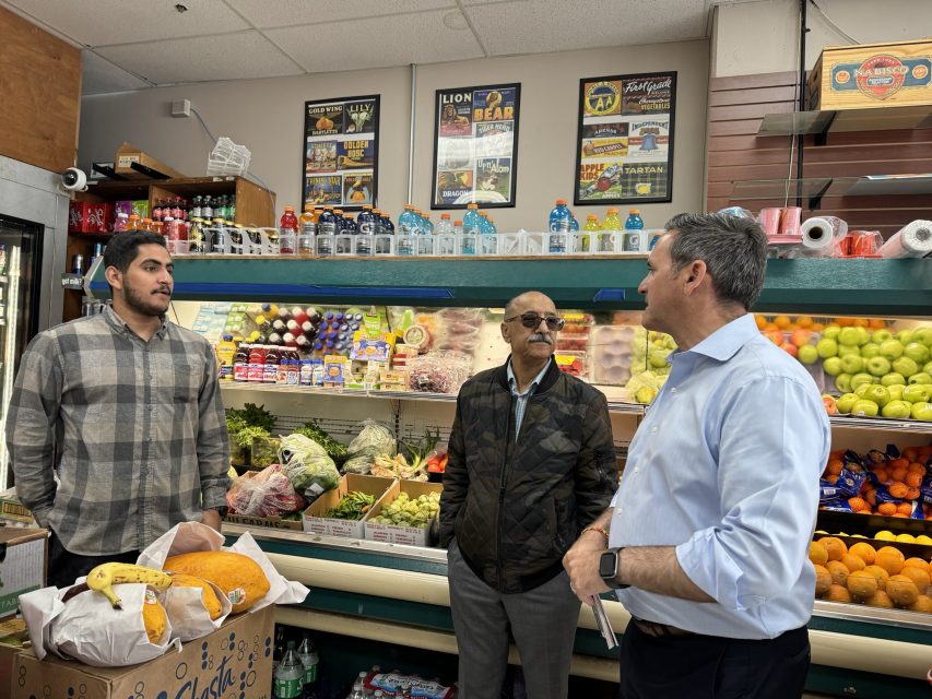 Three men talking inside a grocery store, surrounded by shelves of fruit, vegetables, and various food items.