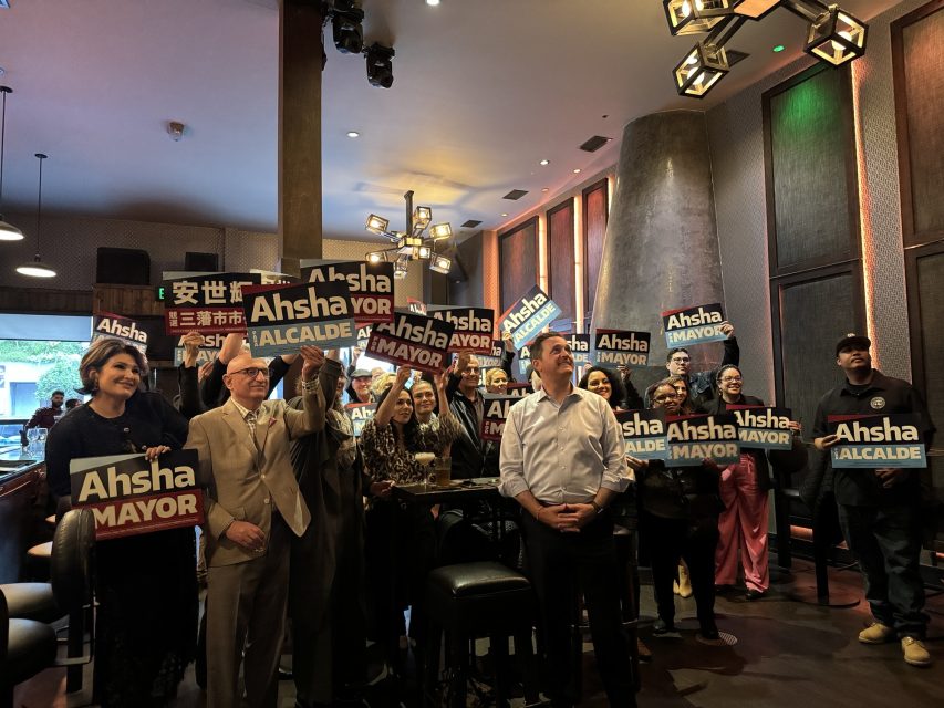 A group of people holds signs that say "Ahsha Mayor" in English, Spanish, and Chinese while gathered indoors in a restaurant-like setting.