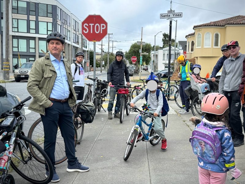 A group of cyclists, including adults and children, stand and sit with their bikes at a street intersection near a stop sign. Several riders wear helmets and casual clothing.