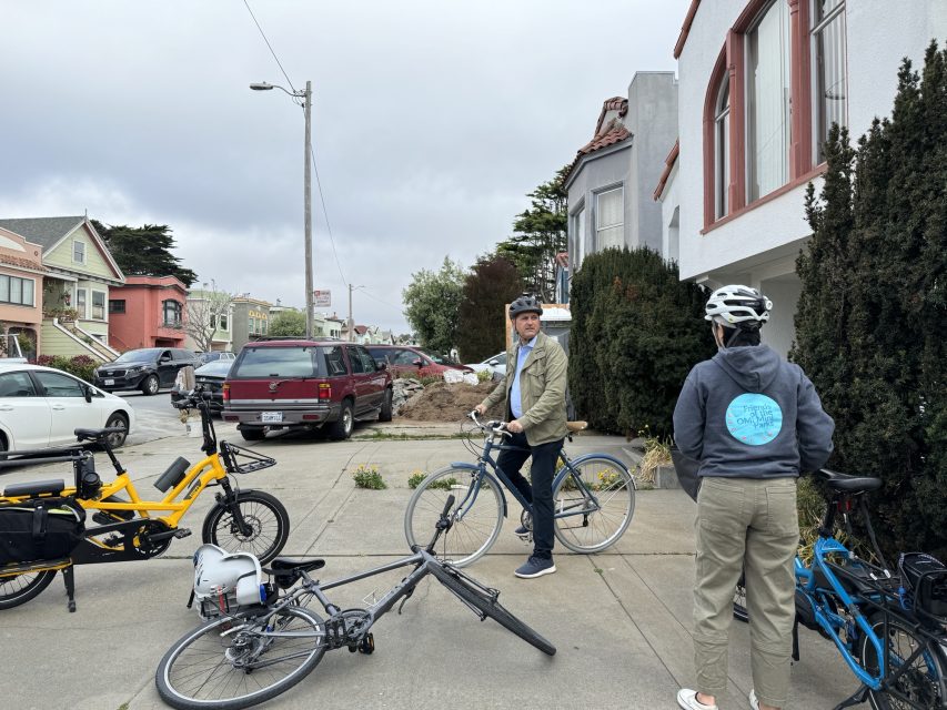 Two people with bicycles are talking on a sidewalk in a residential neighborhood. One person stands, while the other sits on their bike. A bicycle lies on the ground nearby.